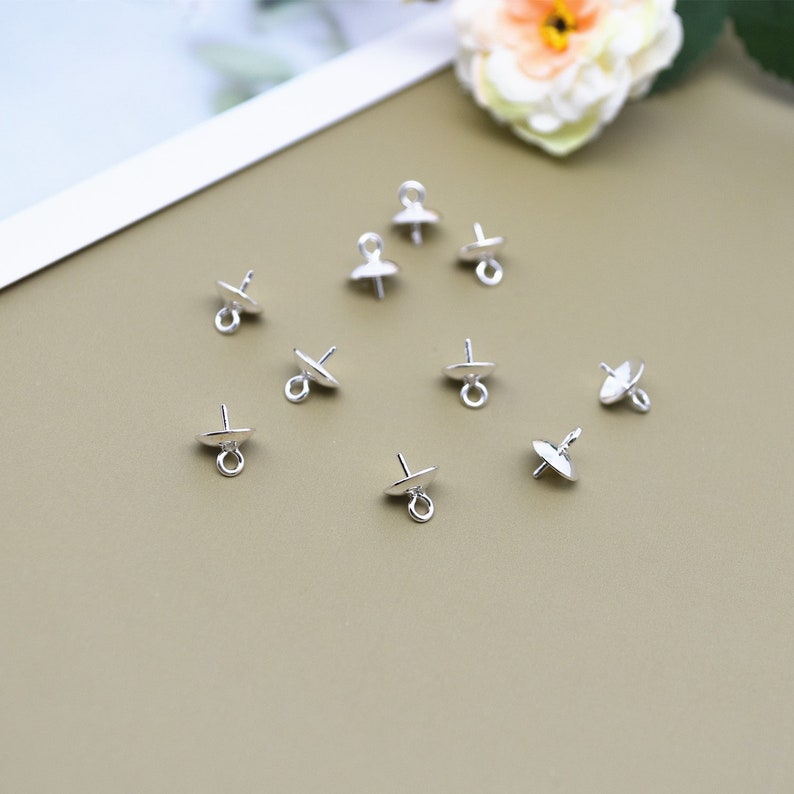 May include: Silver-toned metal bead caps with a loop on the top and a flat, round base. The caps are arranged in a scattered pattern on a brown surface.