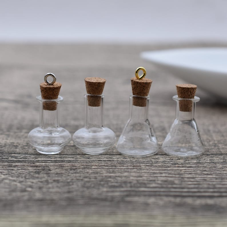 May include: Four small glass bottles with cork stoppers and metal loops. The bottles are clear and have different shapes. They are arranged in a row on a wooden surface.
