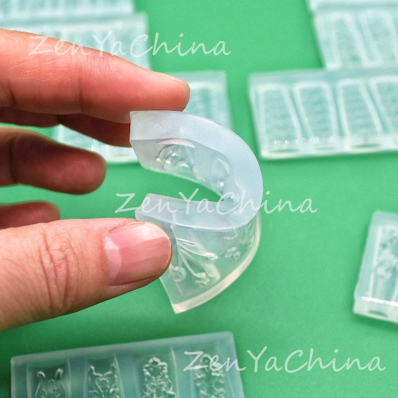 May include: A clear, flexible silicone mold held by a hand, featuring a curved shape with detailed floral and geometric designs. The mold is translucent, with the text "Zen YaChina" visible in the background.