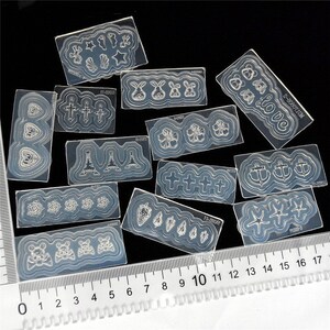 May include: A set of 12 clear nail art stencils with various designs, including hearts, crosses, Eiffel Towers, flowers, anchors, stars, and bunnies. The stencils are arranged on a black surface with a ruler for scale.