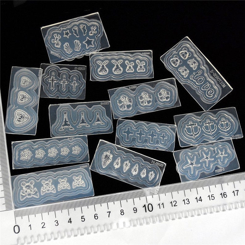 May include: A set of 12 clear nail art stencils with various designs, including hearts, crosses, Eiffel Towers, flowers, anchors, stars, and bunnies. The stencils are arranged on a black surface with a ruler for scale.