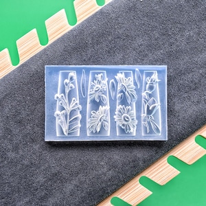 May include: A clear, rectangular mold featuring four floral designs: a sprig of flowers, two sunflowers, and a rose. The mold is set against a gray fabric background, with a green and wood-toned element in the corners.