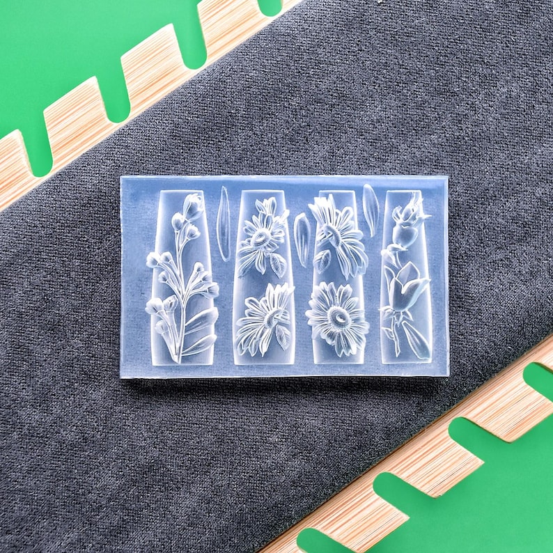 May include: A clear, rectangular mold featuring four floral designs: a sprig of flowers, two sunflowers, and a rose. The mold is set against a gray fabric background, with a green and wood-toned element in the corners.