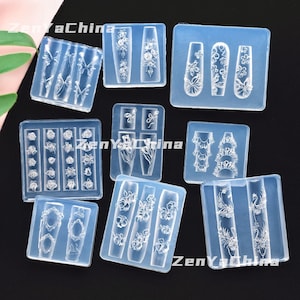 Puede incluir: Una colección de moldes de silicona transparentes para nail art. Los moldes presentan varios diseños, incluyendo flores, mariposas y patrones decorativos. Los moldes están dispuestos sobre una superficie negra.