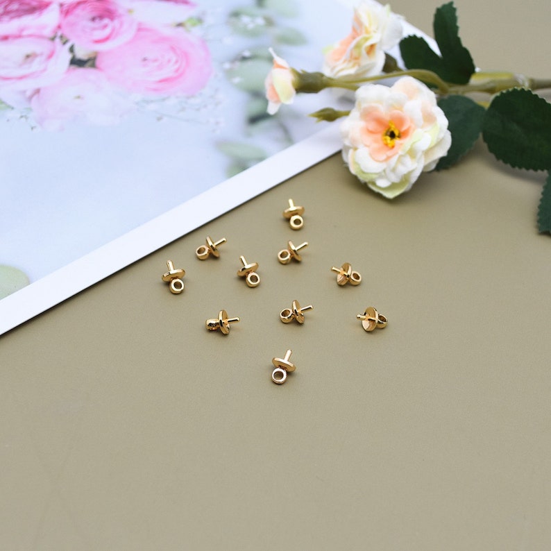 May include: A collection of 10 gold-toned bead tips with a loop on the top. The bead tips are arranged in a circle on a brown surface.