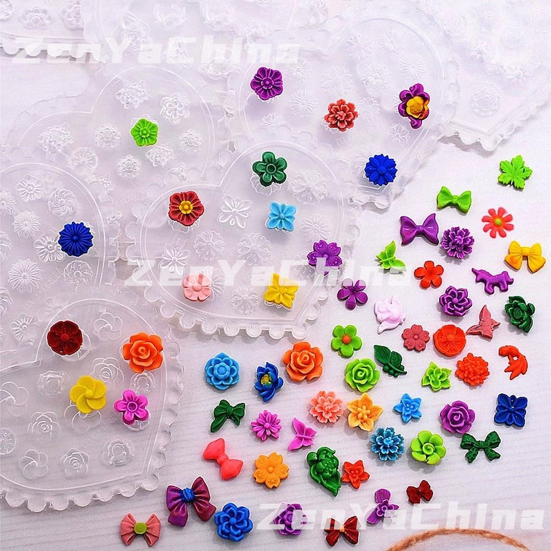 May include: A selection of small, colourful floral and bow-shaped decorations. The collection features various shades of red, orange, yellow, green, blue, and purple. Some are arranged on heart-shaped, lace-edged plastic moulds, while others are scattered.