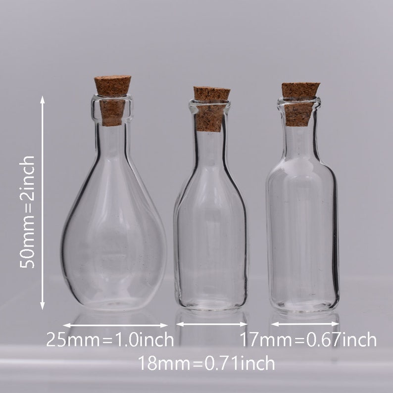 May include: Three clear glass bottles with cork stoppers. The bottles are different sizes. The largest bottle is 2 inches tall and 1 inch wide. The middle bottle is 0.71 inches tall and 0.67 inches wide. The smallest bottle is 0.71 inches tall and 0.67 inches wide.