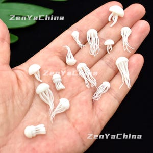 Könnte beinhalten: Eine Sammlung kleiner, weißer Quallenfiguren mit detaillierten Tentakeln und abgerundeten Oberteilen. Die Figuren sind auf einer Hand verteilt, was ihr filigranes Design zeigt. Das Bild ist gut beleuchtet und hebt die Textur und Form jedes Stücks hervor.