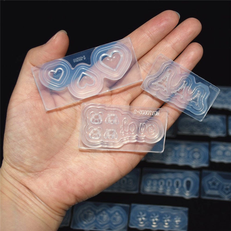 May include: Three clear silicone molds for crafting. The molds feature different designs, including hearts, the word "love", and a stylized Eiffel Tower. The molds are on a hand.