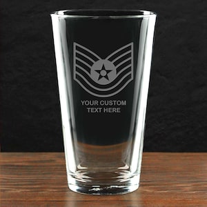 May include: A clear glass pint glass with a military insignia and the text "YOUR CUSTOM TEXT HERE" etched on the front. The glass is set on a wooden surface against a dark background.