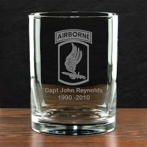 May include: A clear glass tumbler with the word "AIRBORNE" above a military emblem featuring a winged design and a sword. The text "Capt John Reynolds 1990-2010" is etched below the emblem. The glass sits on a wooden surface.
