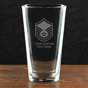May include: A clear glass pint glass with an etched design of a United States Air Force technical sergeant rank insignia. The text "YOUR CUSTOM TEXT HERE" is also etched on the glass.