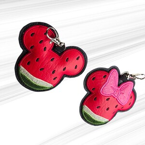 K&ouml;nnte beinhalten: Zwei Schl&uuml;sselanh&auml;nger zum Thema Wassermelone. Einer hat die Form von Mickey Mouses Kopf, der andere hat eine rosa Schleife. Beide sind rot mit schwarzen Konturen, gr&uuml;ner Schale und schwarzen Samendetails. Jeder hat einen silbernen Verschluss.