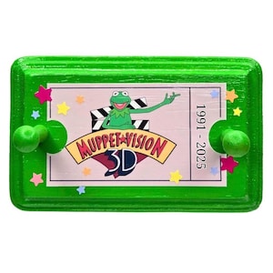 Puede incluir: Placa de madera verde con un diseño de boleto rosa que presenta a la Rana Gustavo y las palabras "Muppet Vision 3D". La placa está decorada con estrellas de colores y perillas verdes. Los años 1991-2025 están impresos a la derecha.