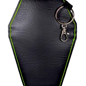 May include: A black coffin-shaped keyring featuring a textured surface and bright neon green stitching along the edges. The item includes a silver-toned metal split ring and a lobster claw clasp attached to the top corner with a black fabric loop.