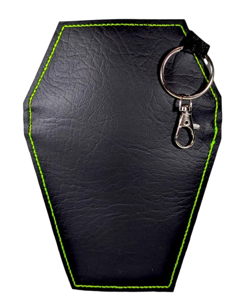 May include: A black coffin-shaped keyring featuring a textured surface and bright neon green stitching along the edges. The item includes a silver-toned metal split ring and a lobster claw clasp attached to the top corner with a black fabric loop.