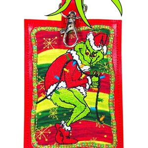 May include: A rectangular red badge holder featuring an embroidered green character wearing a red Santa suit and hat. The character is depicted against a background of red, yellow, and green stripes with gold snowflake accents. A silver metal clasp is attached.