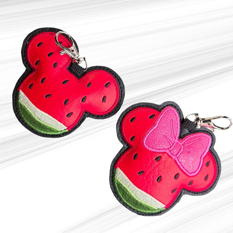 K&ouml;nnte beinhalten: Zwei Schl&uuml;sselanh&auml;nger im Wassermelonen-Design. Einer ist rot mit schwarzen Samen und gr&uuml;ner Schale, schwarz umrandet. Der andere hat eine rosa Schleife. Beide haben silberne Verschl&uuml;sse.