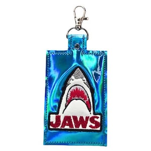 Puede incluir: Un porta credenciales rectangular de color azul iridiscente con un parche bordado de un tiburón con la boca abierta y el texto rojo JAWS debajo. El soporte incluye un mosquetón de metal plateado sujeto en la parte superior.