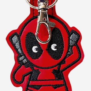 以下が含まれることがあります： 丸い頭、黒いアイマスク、白い瞳を持つ漫画のキャラクターが刺繍された赤いキーホルダー。キャラクターは頭の後ろに2本の灰色の剣を持っています。上部の赤い布ループには、銀色の金属製ロブスタークラスプが取り付けられています。