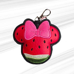 K&ouml;nnte beinhalten: Ein roter und schwarzer Schl&uuml;sselanh&auml;nger im Wassermelonen-Design mit einer rosa Schleife. Der Schl&uuml;sselanh&auml;nger hat die Form eines M&auml;usekopfes mit einem Wassermelonen-Design, einschlie&szlig;lich schwarzer Samen und einer gr&uuml;n-wei&szlig;en Schale. Er hat einen silberfarbenen Verschluss.