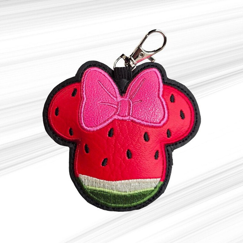 K&ouml;nnte beinhalten: Ein roter und schwarzer Schl&uuml;sselanh&auml;nger im Wassermelonen-Design mit einer rosa Schleife. Der Schl&uuml;sselanh&auml;nger hat die Form eines M&auml;usekopfes mit einem Wassermelonen-Design, einschlie&szlig;lich schwarzer Samen und einer gr&uuml;n-wei&szlig;en Schale. Er hat einen silberfarbenen Verschluss.