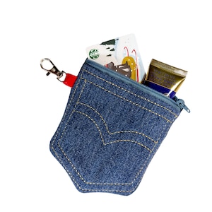 May include: A small blue denim pouch with a zipper closure and a red and silver key chain. The pouch has a pocket on the front and is filled with a gift card and a tube of hand cream.