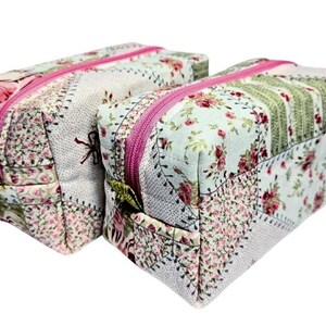 Könnte beinhalten: Zwei rechteckige Kosmetiktaschen mit floralen Patchworkmustern. Die Taschen haben rosa Reißverschlüsse und sind aus Stoff mit einer Mischung aus floralen Drucken in Rosa, Grün und Weiß gefertigt. Die Taschen stehen nebeneinander.