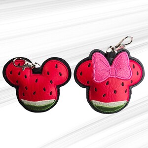 K&ouml;nnte beinhalten: Zwei Schl&uuml;sselanh&auml;nger zum Thema Wassermelone. Einer hat die Form eines Mickey Mouse-Kopfes, der andere hat eine rosa Schleife. Beide sind rot mit schwarzen Samen und einer gr&uuml;nen Schale. Jeder Schl&uuml;sselanh&auml;nger hat einen silbernen Verschluss.