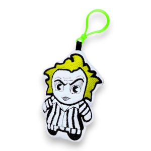 Op de afbeelding: Een pluche sleutelhanger met een stripfiguur met geel haar en een zwart-wit gestreept outfit. Het personage heeft een strenge uitdrukking en is bevestigd aan een groene lus.
