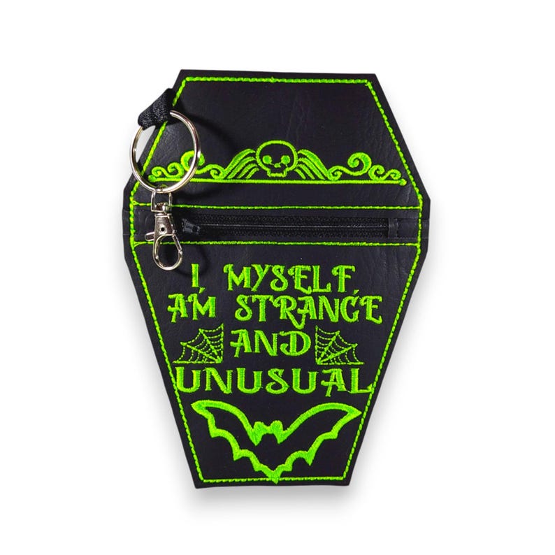 May include: A black coffin-shaped pouch with a silver metal keyring. The pouch features bright green embroidery including a skull, spiderwebs, a bat, and the text "I, MYSELF, AM STRANGE AND UNUSUAL" with a horizontal zippered closure across the top.