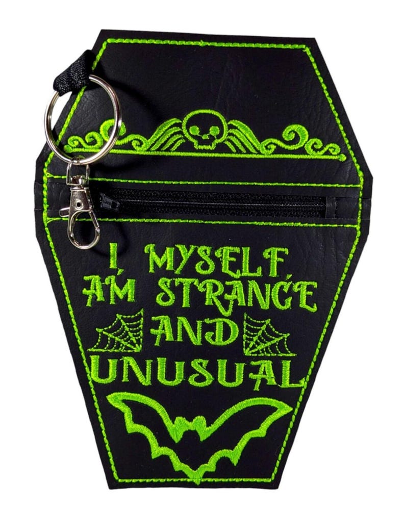 May include: A black coffin-shaped pouch with bright green embroidery featuring a skull, spiderwebs, a bat, and the text I, MYSELF, AM STRANGE AND UNUSUAL. The pouch includes a black zip closure and a silver metal keyring with a lobster claw clasp.