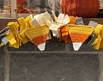 May include: A festive garland featuring fabric pieces in yellow, orange, gray, and white, resembling candy corn. The garland is strung along a wooden beam, perfect for autumn or Halloween decor.