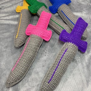 May include: A collection of handmade crochet toy swords in various colors. The swords feature gray blades with colored accents and brightly colored handles in pink, green, blue, yellow, and purple. The toys are soft and likely for children.