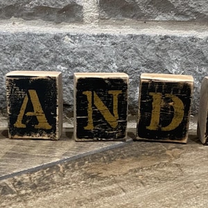 May include: Five wooden blocks spelling out "VANDY" in gold letters against a black background. The blocks have a distressed, vintage look, with visible wood grain and a weathered appearance. The blocks are arranged on a wooden surface.