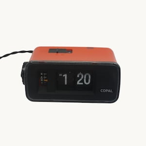 Op de afbeelding: Een oranje en zwarte vintage Copal klok met "1:20 AM". De klok heeft een rechthoekig ontwerp met een zwart digitaal display en een kleine draaiknop aan de zijkant. Het woord "Copal" staat rechtsonder.