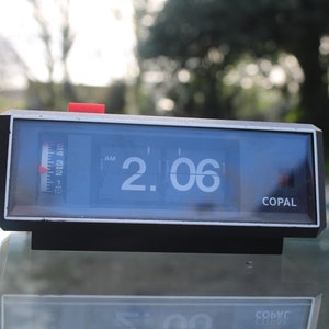 May include: A black flip clock with a white face displaying 2:06 AM. The clock has a red button on the top and the word "COPAL" printed on the bottom.
