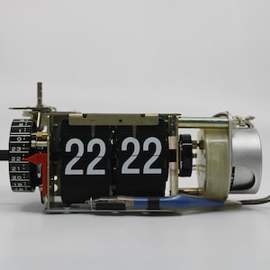 May include: A vintage flip clock displaying the time 22:22. The clock has a black face with white numerals and is housed in a metal frame. The clock mechanism is visible, with wires and gears.
