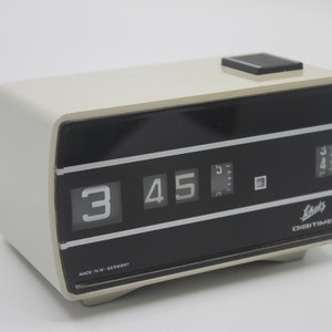May include: A white plastic digital clock with black flip numbers displaying 3:45. The clock has a black face with the text "Leitz Digitime" and "Made in W.-Germany".
