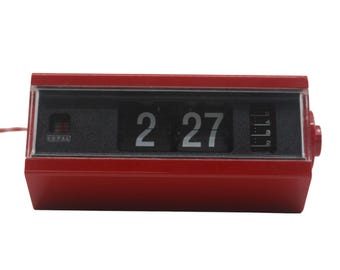 May include: A vintage red flip clock displaying the time 2:27. The clock has a rectangular shape with a black display area and a red casing. The brand name "COPAL" is visible on the left side of the display.