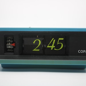Può includere: Un orologio digitale blu con un quadrante nero e numeri gialli che mostrano le 2:45. L'orologio ha una copertura in plastica trasparente e la parola "COPAL" è stampata in basso a destra.