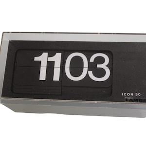 May include: A black flip clock with white numbers displaying 11:03. The clock is in a clear plastic case and has the text "ICON 30 LAMBERT" on the bottom.