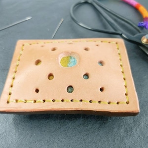 May include: A light brown leather craft with a rectangular shape, featuring a series of small, evenly spaced holes. The edges are stitched with yellow thread. The center has a circular design with blue and yellow accents. Crafting tools are in the background.