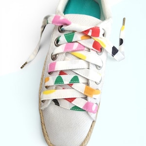 May include: A white canvas sneaker featuring a jute-wrapped sole and a teal interior lining. The shoe is laced with wide, flat fabric laces decorated with a colorful pattern of pink, red, yellow, green, and black abstract shapes and dots.
