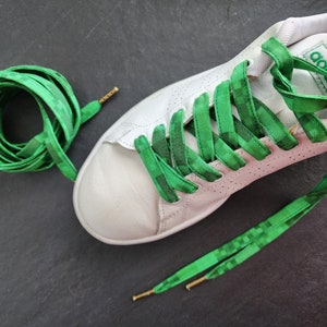 May include: White sneakers with green patterned shoelaces. The shoelaces are tied in a criss-cross pattern and the laces are also shown lying on a dark gray surface.