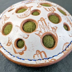May include: A white, round, decorative object with brown etched designs and green filling. The object has a blue stitched border and is decorated with bird and hand motifs. The object has several circular openings.