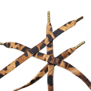 May include: Shoelaces with a leopard print pattern. The laces are brown and black with gold-colored aglets. The laces are arranged in a crisscross pattern against a white background.