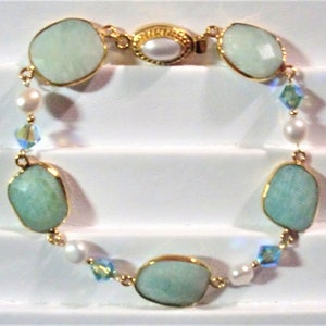 May include: A gold-toned bracelet featuring large, light blue-green gemstone beads with gold edging. The bracelet also includes small white pearls and iridescent crystal accents, connected by gold links.