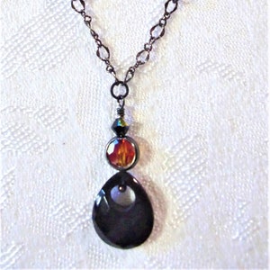 Puede incluir: Un collar de cadena negra con un colgante en forma de lágrima. El colgante es negro con un anillo plateado alrededor de la parte superior. El collar tiene dos cuentas de vidrio rojo y naranja con acentos negros.