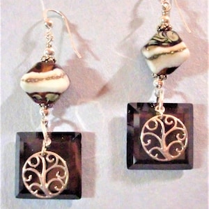 May include: A pair of dangle earrings with silver hooks. Each earring has a square, dark brown, faceted bead with a silver tree of life charm. Above, a decorative bead with brown, white, and green accents.